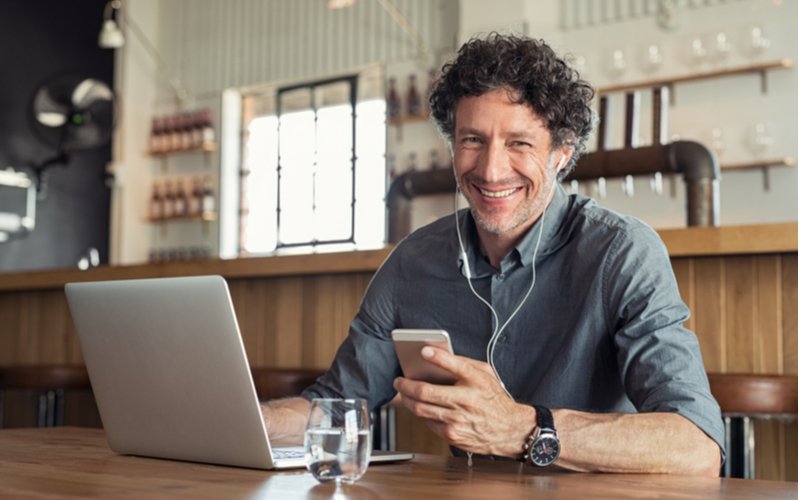 A middle-aged man using his phone and laptop in a cafe, smiling at the camera.