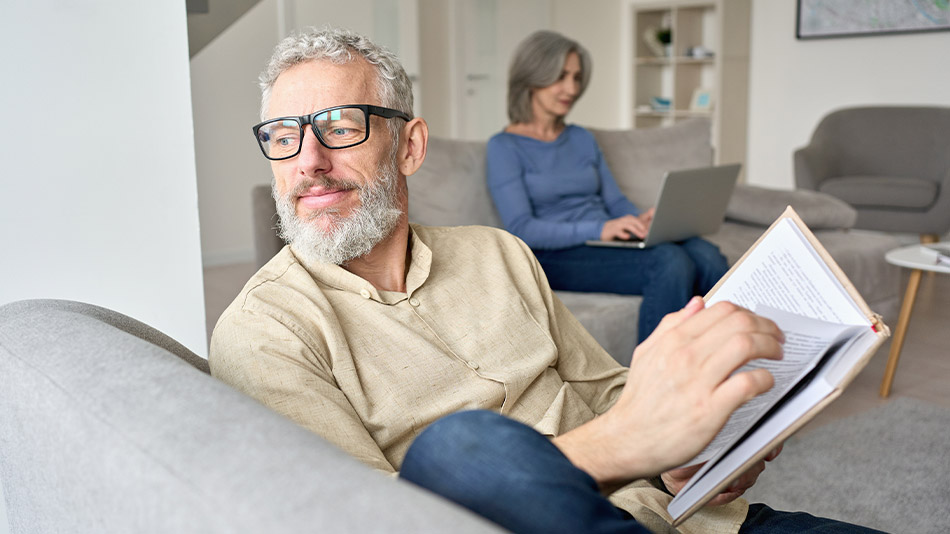 A senior man reading on a sofa at home while wearing glasses