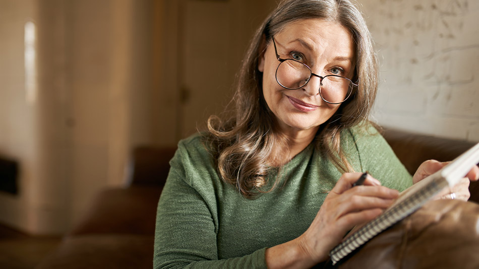 A senior woman comparing lens replacement and reading glasses, taking notes with pen and paper