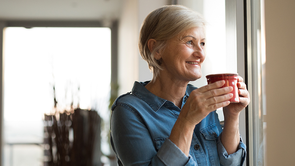 A senior woman holding a red coffee cup, looking out of her window and smiling.