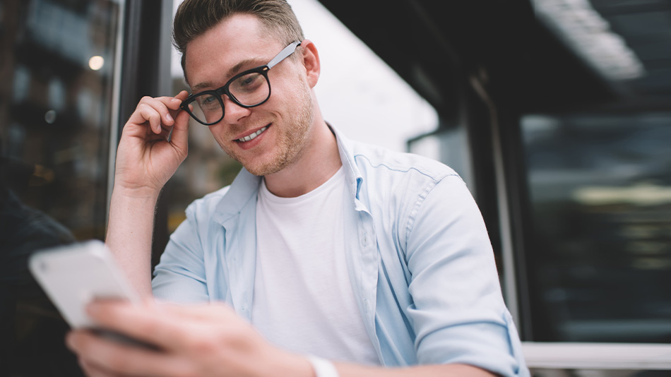 A young man touching his glasses while using his phone.