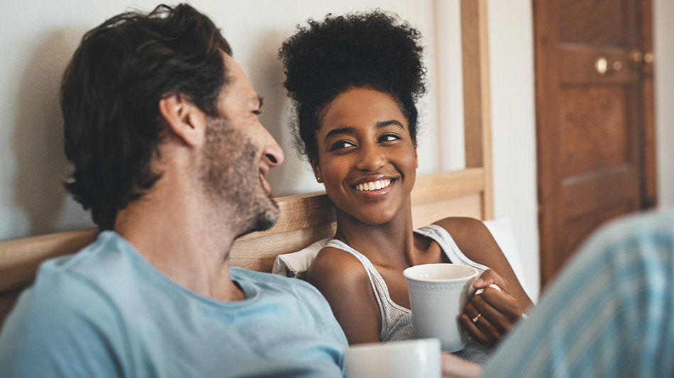 A young couple with a cup of coffee, smiling at each other on the bed in the morning.