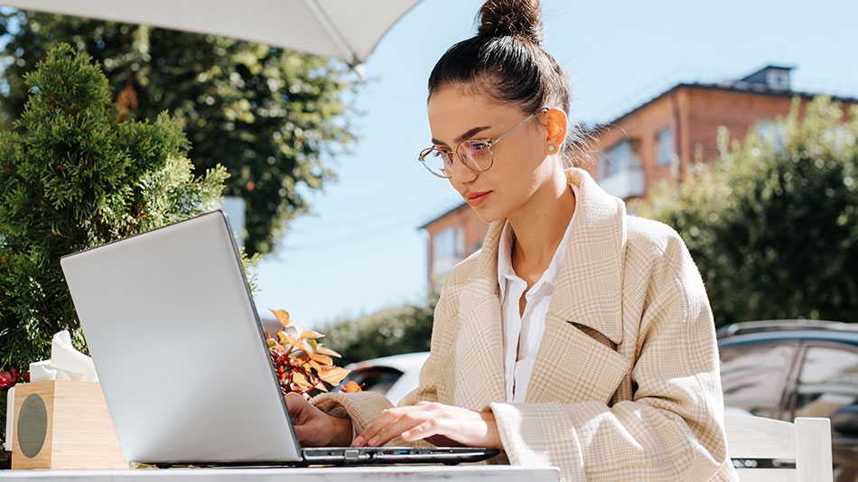 A woman with glasses sitting outdoors on a sunny day, looking at her laptop.