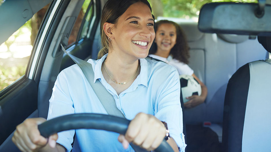 A young mother smiling and driving with her young daughter in the back seat.
