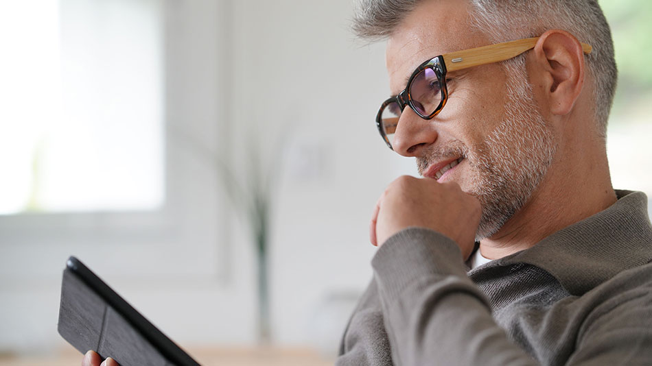 A senior man sits on a couch reading on his iPad.