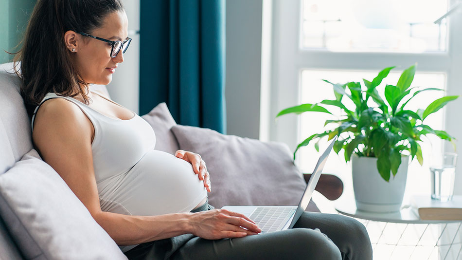 A pregnant woman with glasses reads on her laptop at home.