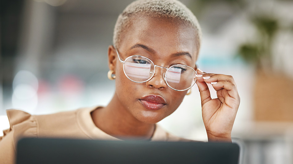 A young black woman touches her glasses while reading on her laptop.