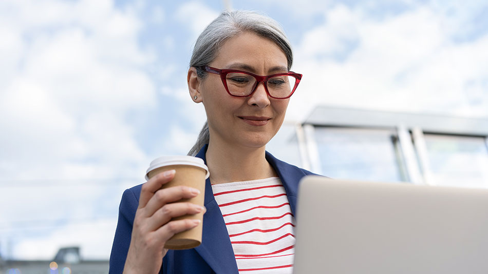 A senior woman with glasses holds a coffee while working on her laptop.