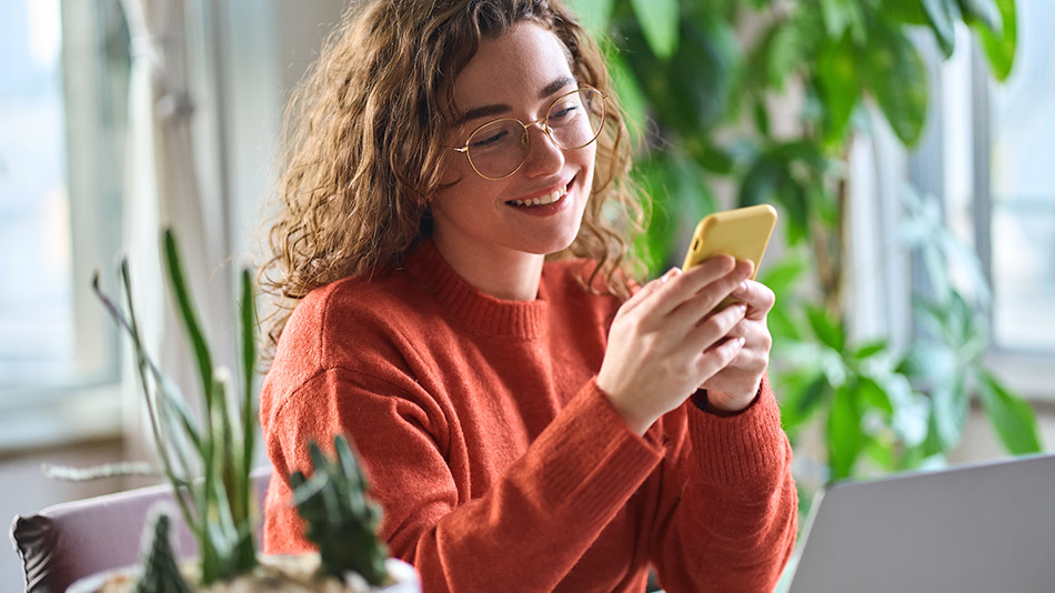 A woman with curly hair and glasses smiles while using her phone.