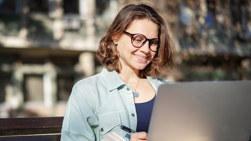 A young woman with glasses sits outside using her laptop.