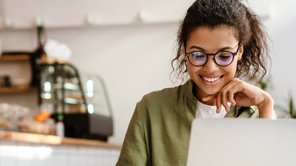 A stylish young woman with curly hair and glasses sits in a sunny coffee shop, wearing a green t-shirt and using a laptop.