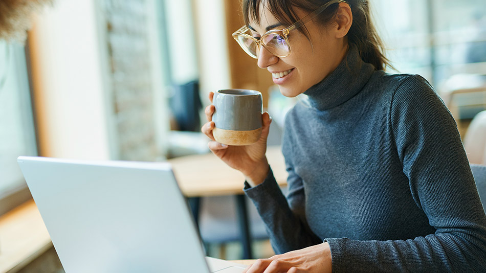 "A young woman with glasses holds a cup of coffee while looking at her laptop."