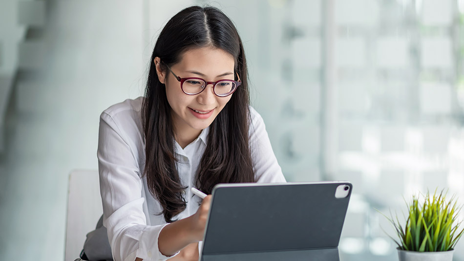 A middle-aged Asian woman with glasses is looking at her iPad in an office.