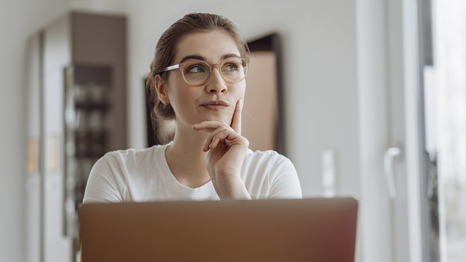 A thoughtful young blonde woman wearing glasses gazes upward while pondering, with an open laptop in front of her.