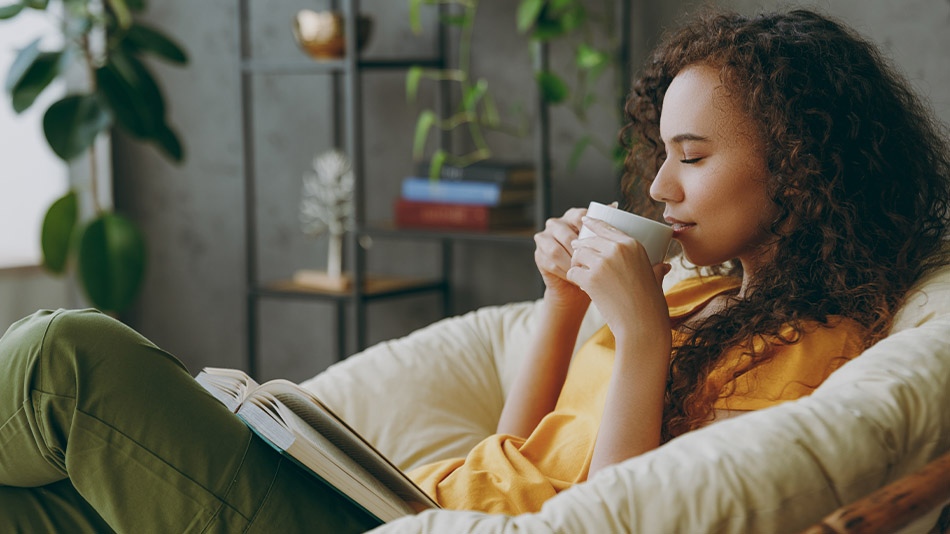 A woman with long hair sits on a sofa at home, holding a cup of coffee and engrossed in a book.