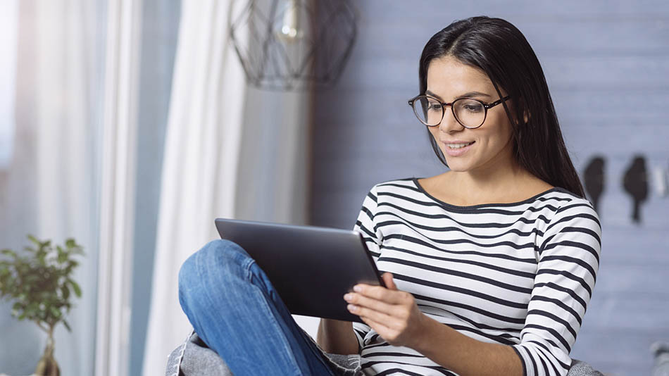 A young woman with long hair and glasses uses her iPad while lounging on a sofa.