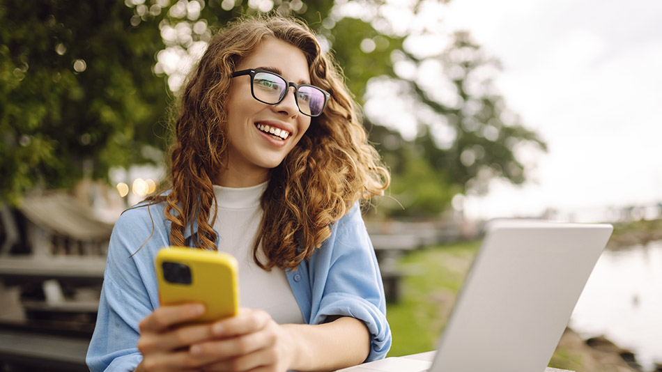 A young woman with glasses and curly hair sitting at a picnic table, holding a phone and using her laptop.