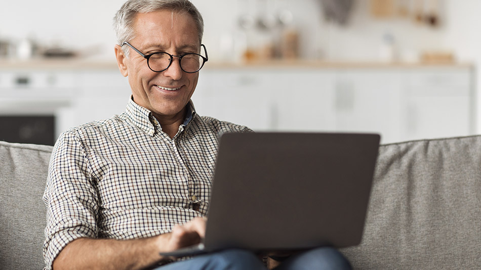 A senior man in a plaid shirt, smiling as he sits on a couch and uses a laptop.