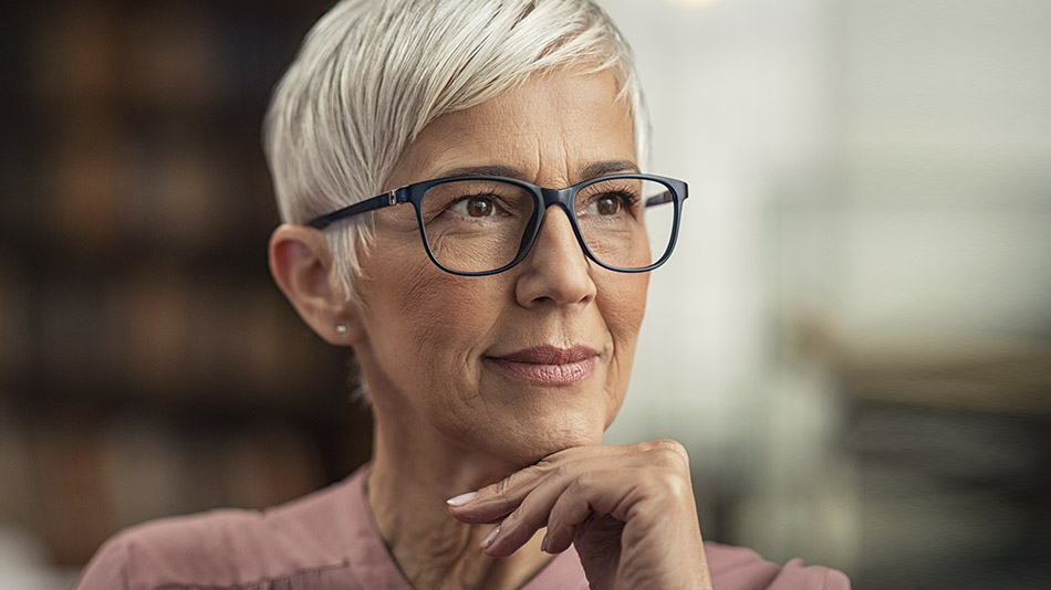 A close-up of a mature woman with glasses looking curiously away from the camera with a slight smile.