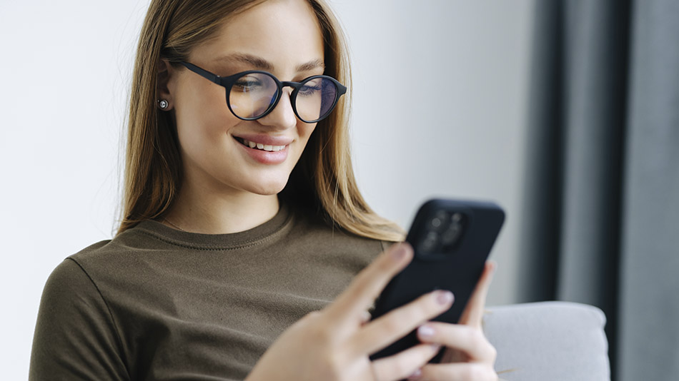 A cheerful young woman with long hair and glasses, smiles as she looks at her phone.
