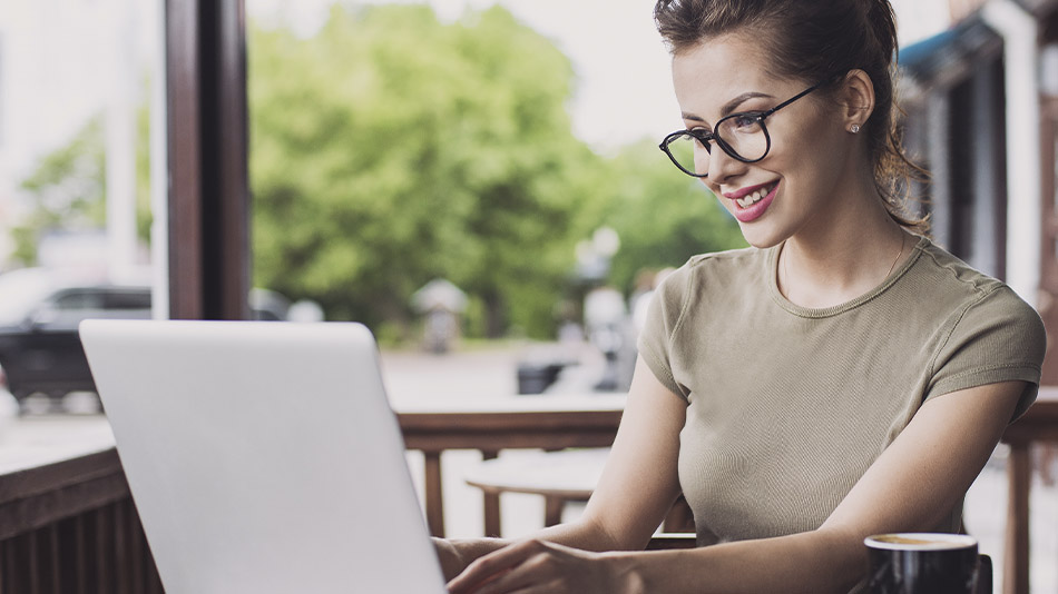 A young woman with glasses sitting on an outdoor patio, smiling at her laptop.