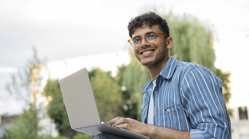 A handsome young man with glasses smiling expectantly while using his laptop outside.