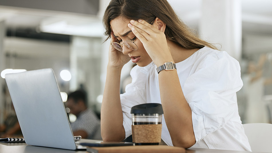 A stressed young woman in a café, sitting in front of her laptop with her hands on her temples.