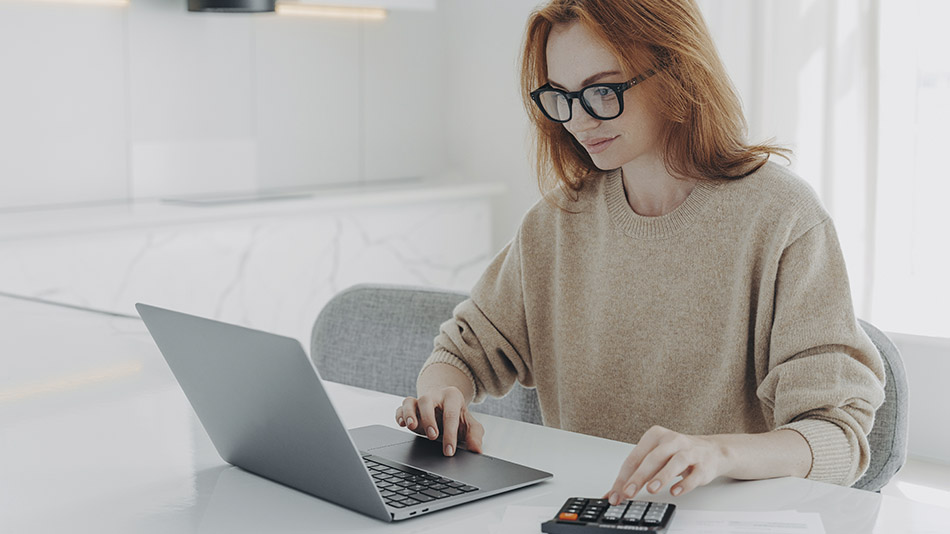 A young woman with glasses sitting at a counter, using a laptop and calculator.