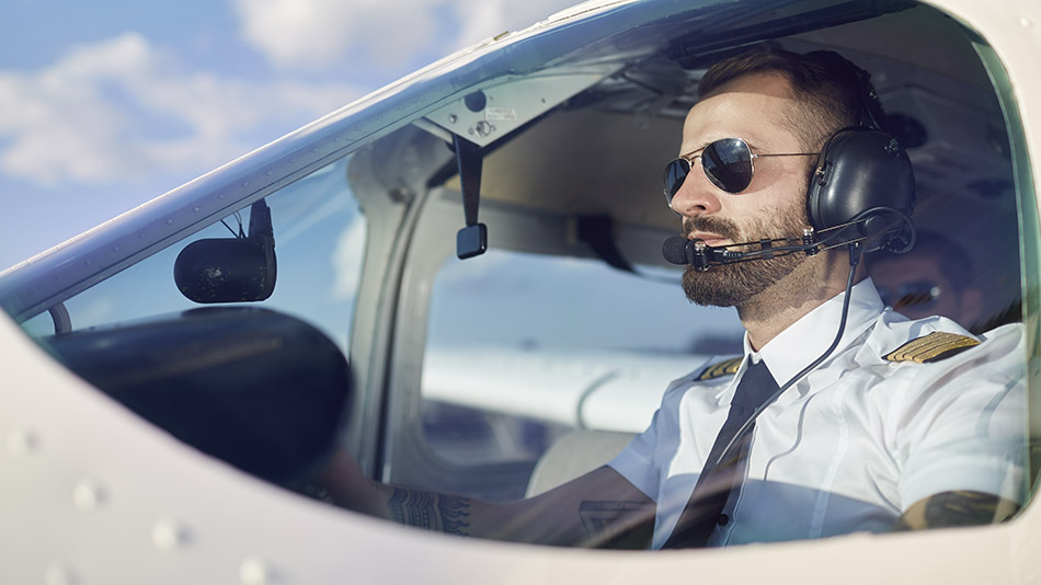A focused young male airplane pilot sitting in the cockpit, wearing sunglasses and a headset.