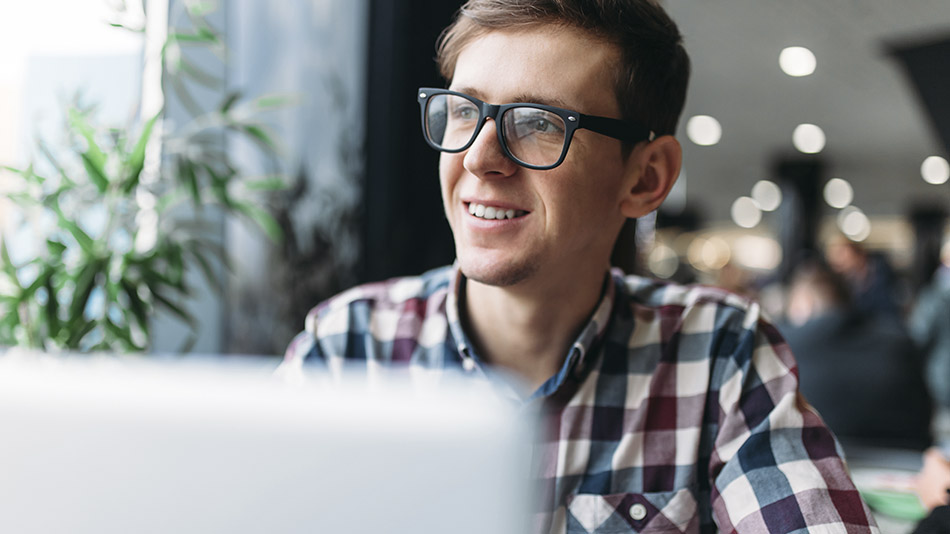 A smiling young man wearing glasses and a plaid shirt, using his laptop in a café.