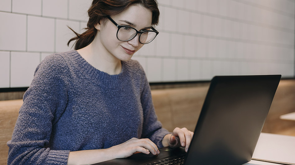A beautiful young woman with glasses smiles gently at her laptop while in a café.