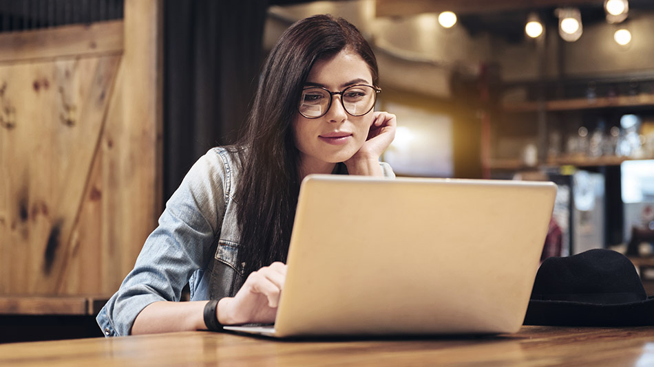 A young woman with glasses is sitting in a wooden café, pondering with an open laptop in front of her.