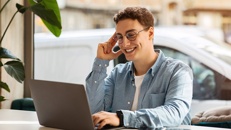 A young man with glasses and curly hair smiling happily at his laptop while sitting in a café.