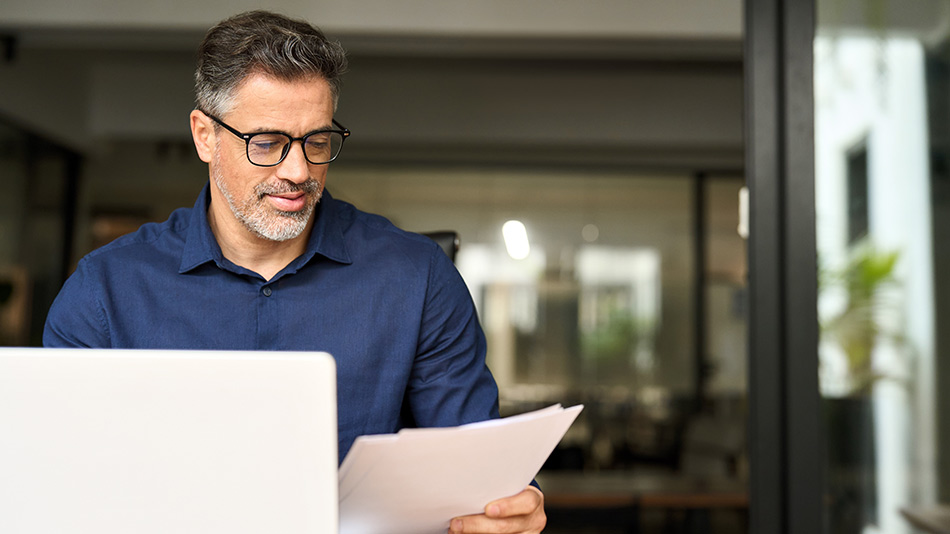 A middle-aged man with glasses reviewing papers in front of his laptop at a desk.