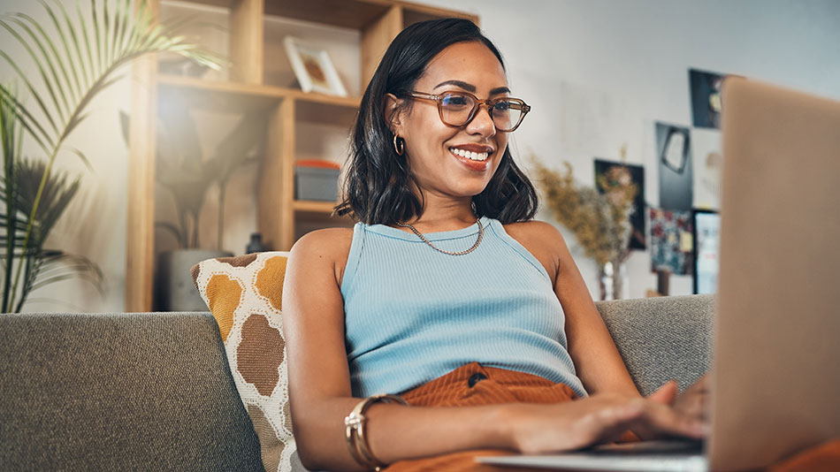 A young woman with glasses using her laptop while lounging on her couch.