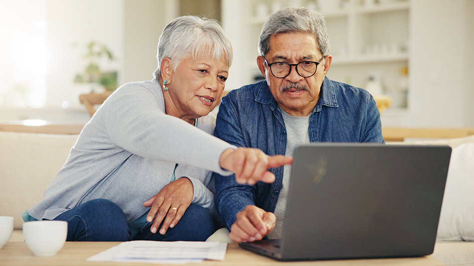 A senior couple sitting on a couch together, looking at a laptop.